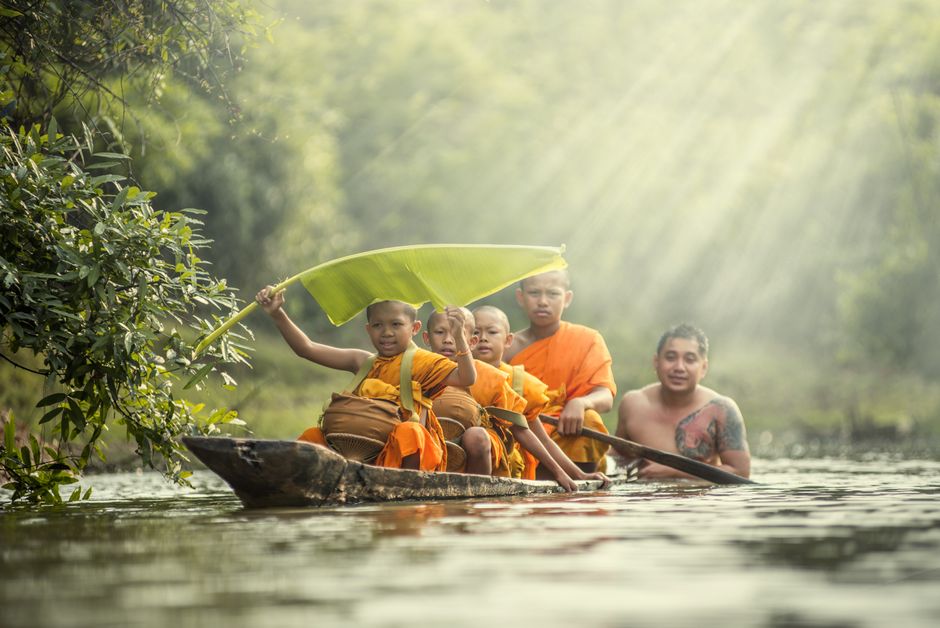 A group of young monks and a man paddling a wooden boat on a serene river, with sunrays filtering through trees.