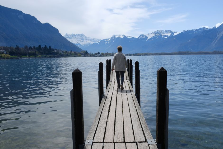 A lone figure walks on a dock with Swiss mountains as a backdrop.