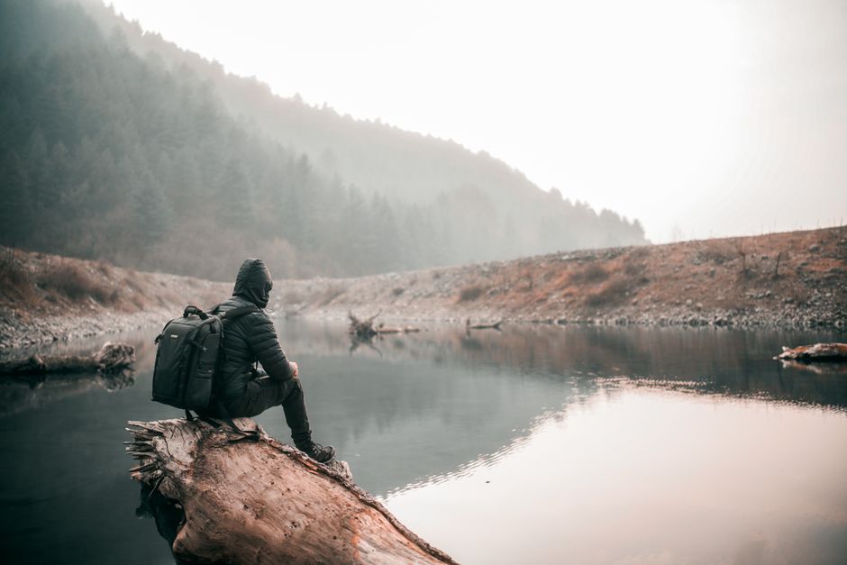 A lone hiker sits on a log by a misty lake, surrounded by foggy trees, exuding tranquility and adventure.