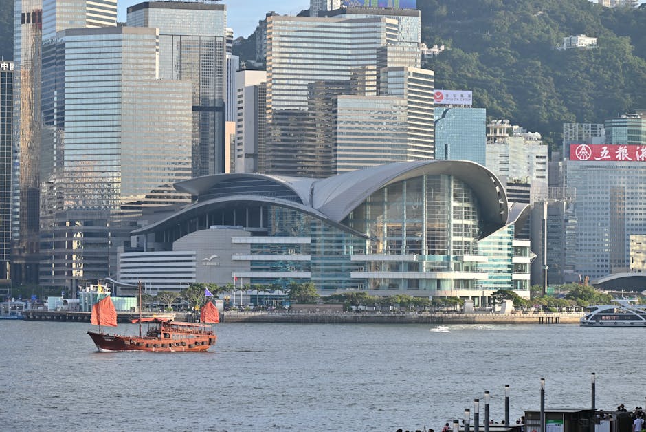 A traditional red-sailed junk boat sails in front of Hong Kong's modern skyline and convention center.
