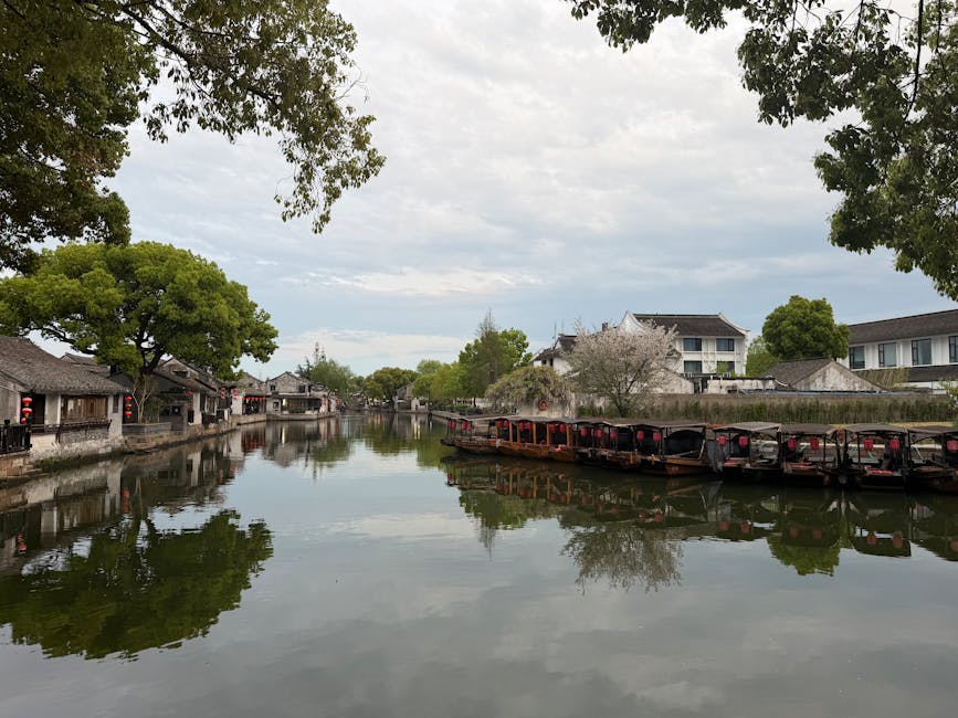 Tranquil view of a historic Chinese village with boats along a serene waterway under a cloudy sky.