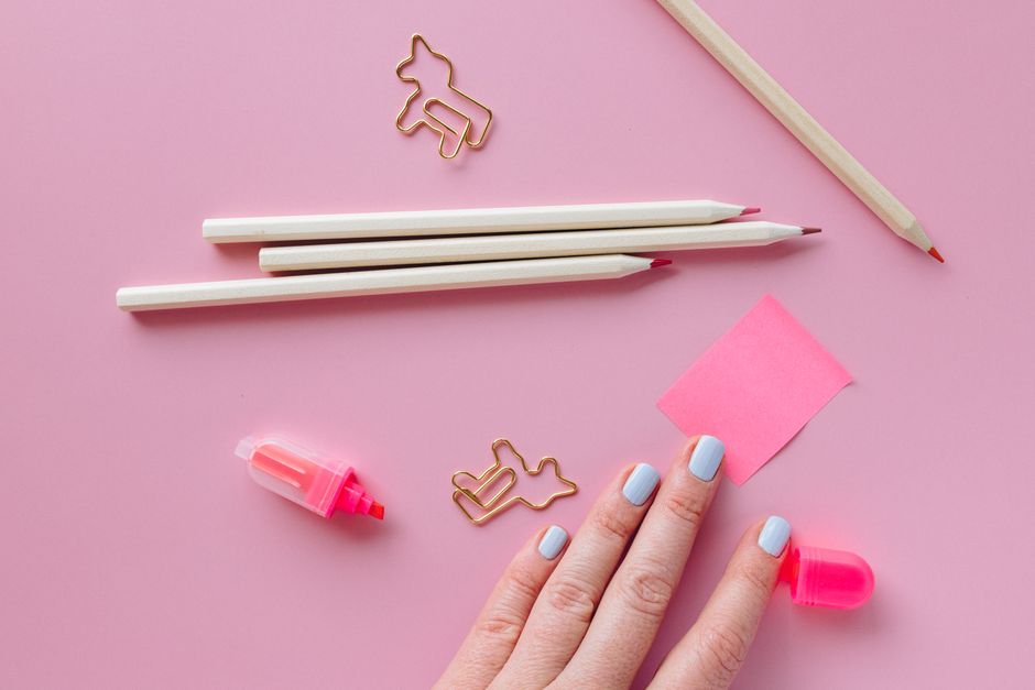 Top view of colorful stationery items on a pink background with a hand featuring painted nails.
