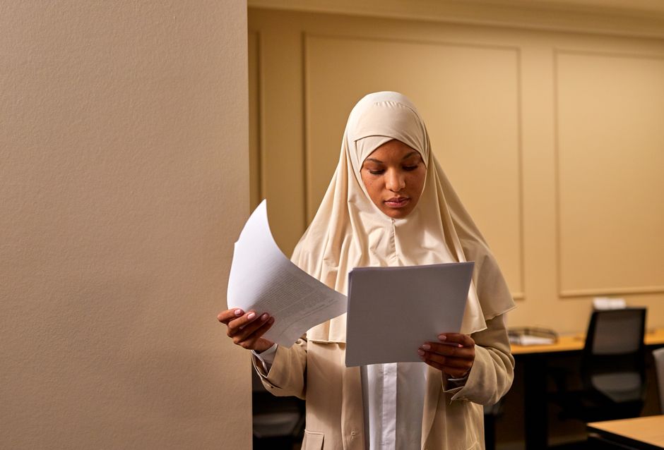 A professional woman in a hijab reviews documents in an office setting.