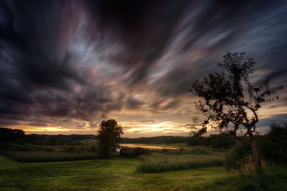 A dramatic sunset casts vibrant colors over a serene rural landscape with dark clouds.