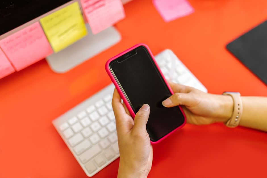 Two hands holding a smartphone above a colorful desk with sticky notes and a keyboard.
