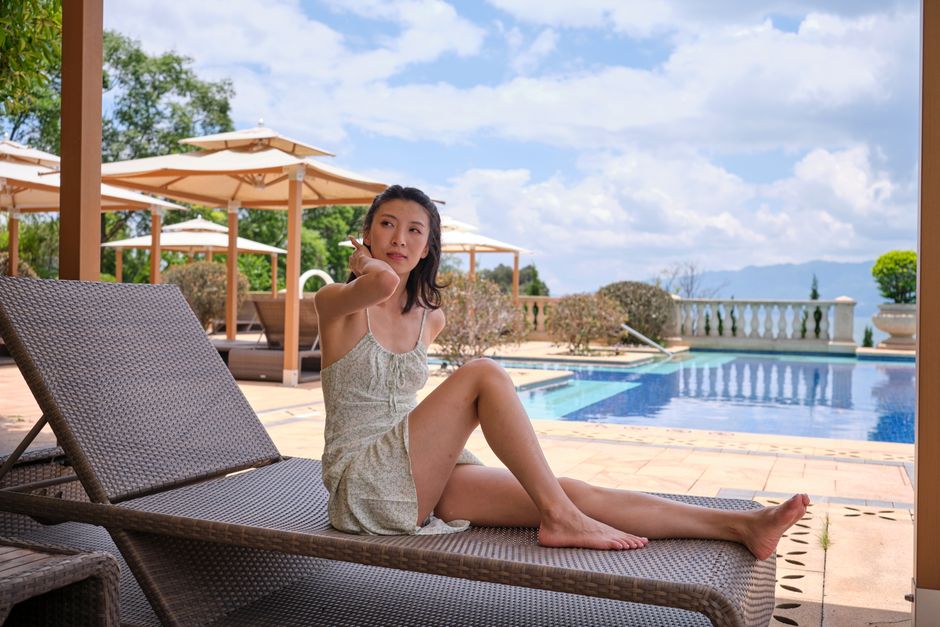 An adult woman in a summer dress relaxes on a lounge chair by a resort pool under a sunny sky.