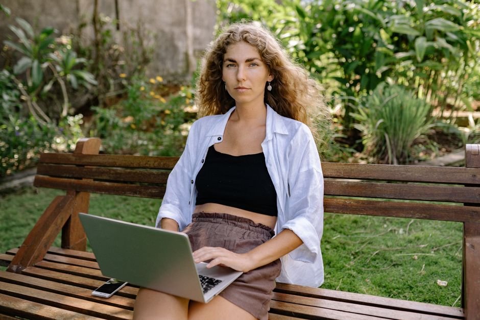 Young woman with laptop sitting outdoors on a wooden bench in a lush garden.