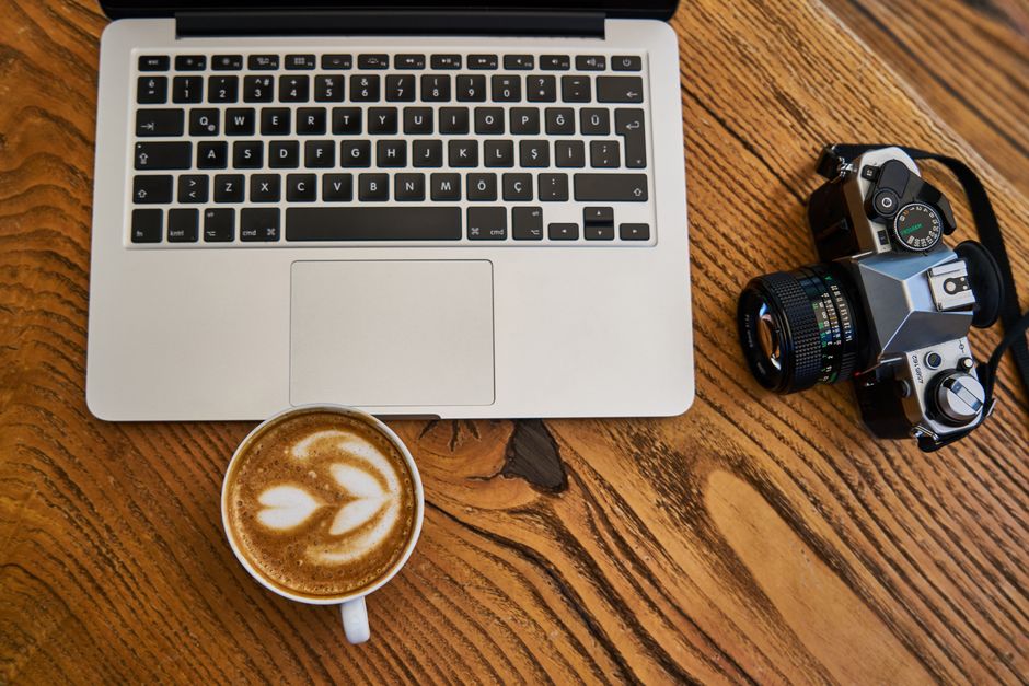 Flat lay of a laptop, cappuccino, and camera on a wooden surface, ideal for work-from-home themes.