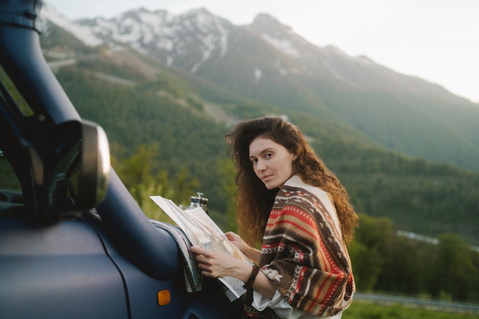 A woman with long hair holding a map by a car in a scenic mountainous landscape.