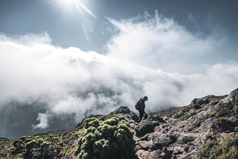 A lone hiker ascends a rocky mountain amidst clouds under a bright sun.