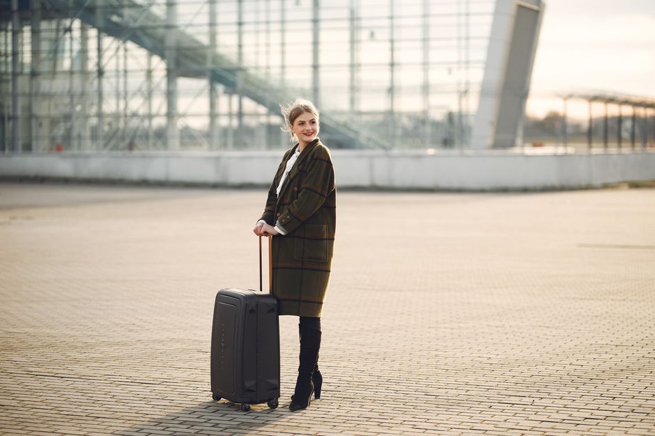 Full length of young joyful woman in outwear and black high heeled boots with luggage standing against glass wall of airport building and looking away