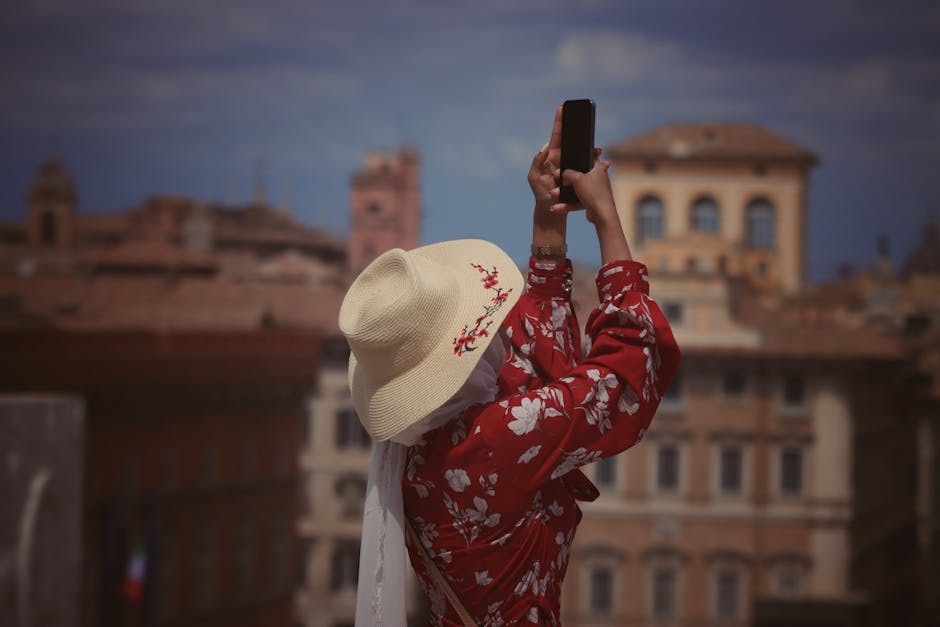 A woman wearing a floral dress and hat takes a picture with a smartphone in a European city.