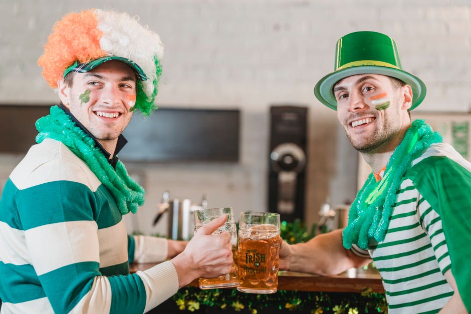 Happy young men in St Patricks day clothes clinking glasses of cold delicious beer at home on blurred background