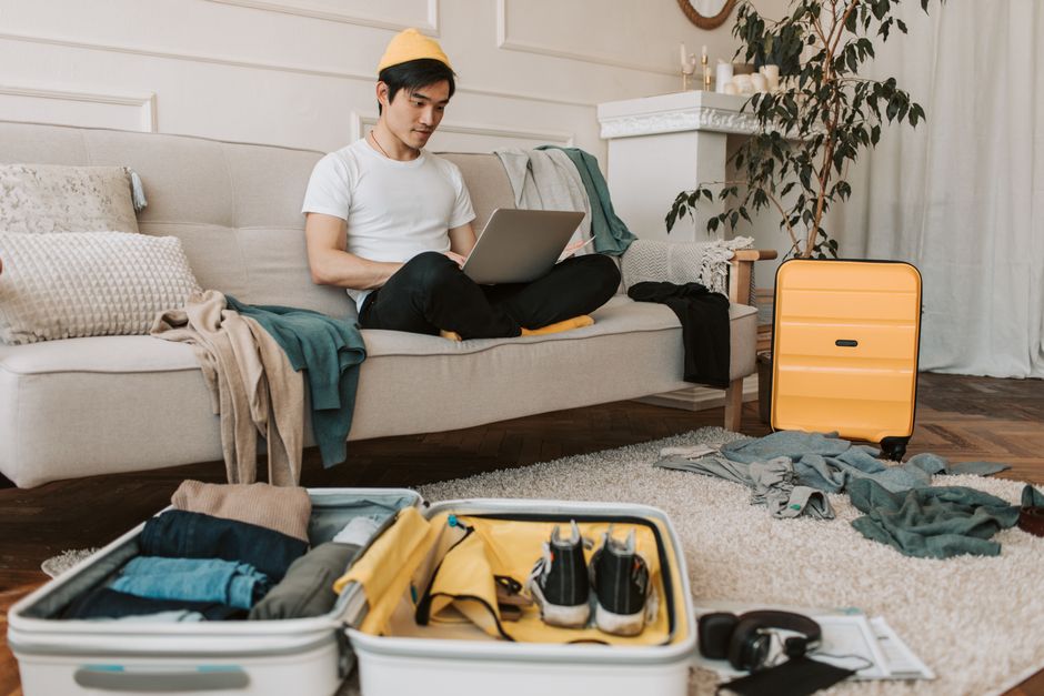 A young man in yellow accessories works on a laptop among unpacked luggage at home.