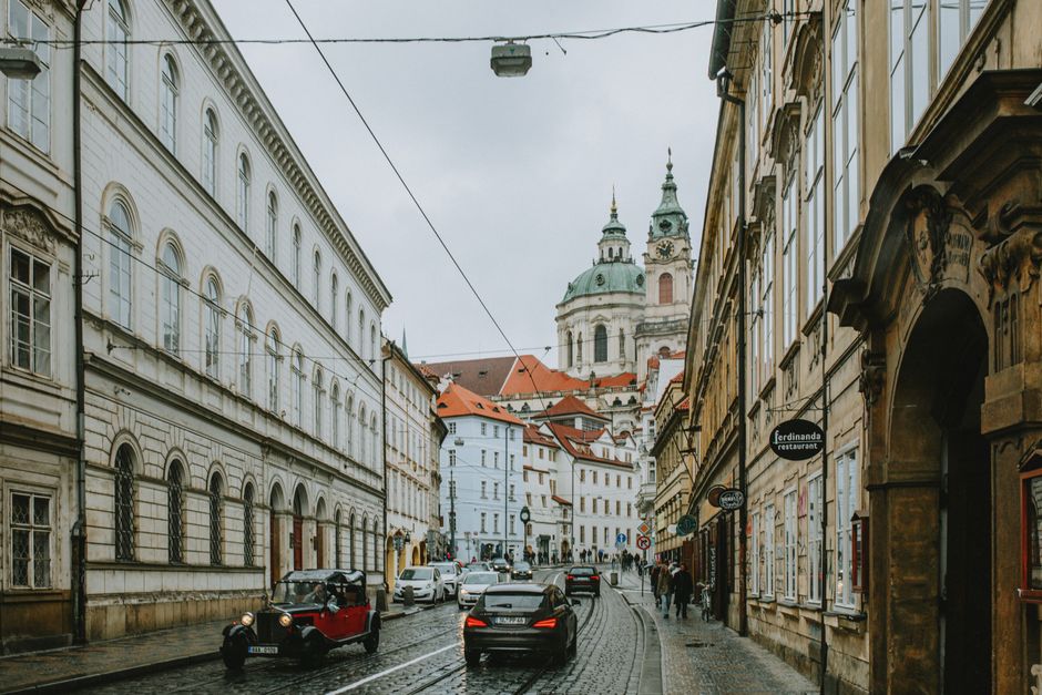 Charming street view showcasing historic architecture in Prague, features classic cars and bustling city life.