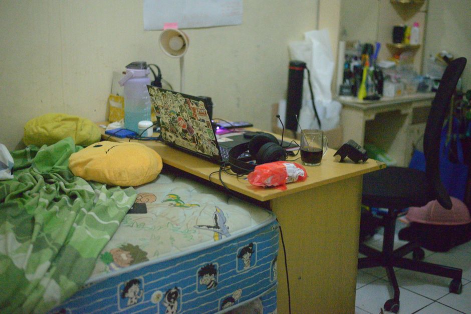 A cluttered workspace with a laptop, headset, and bed in a cozy room setting.