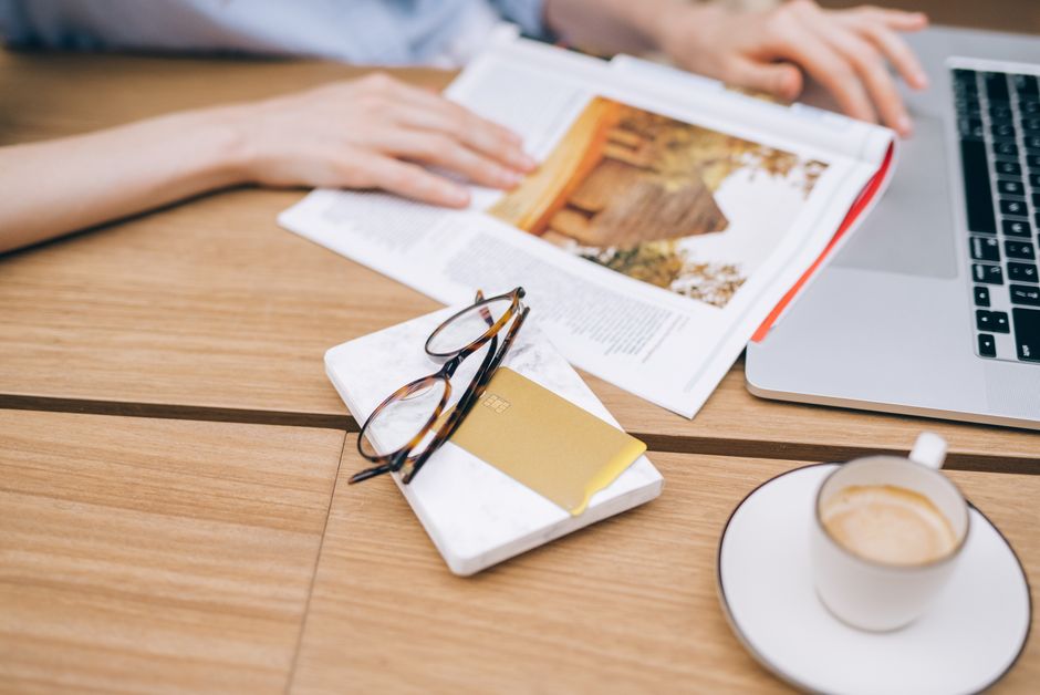 A warm workspace featuring a laptop, coffee cup, magazine, and eyeglasses on a wooden table.