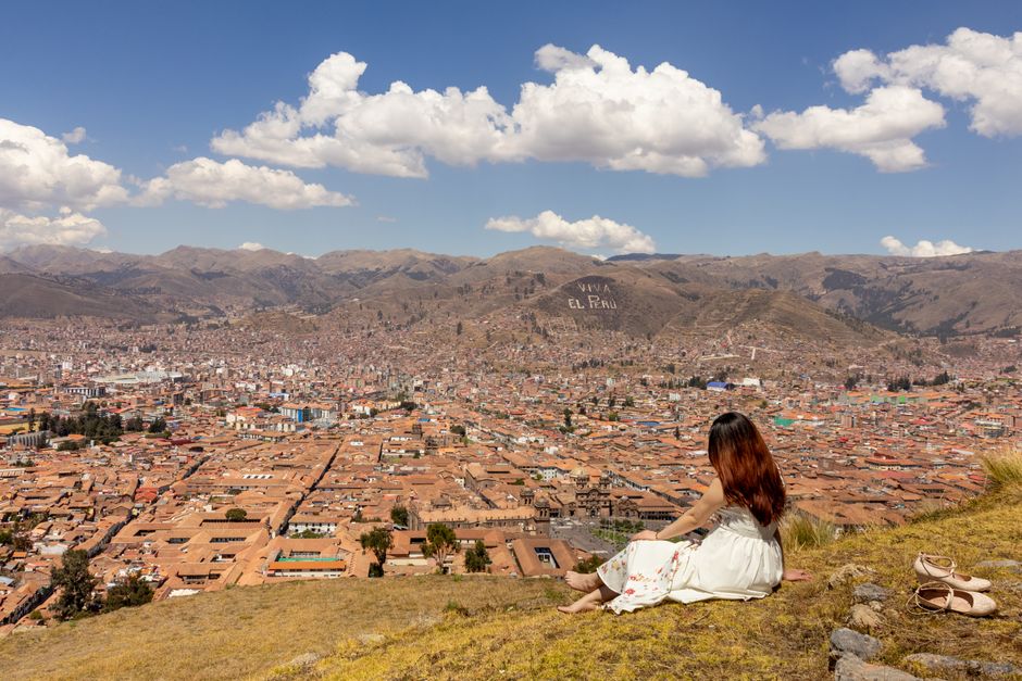 A woman sits atop a hill, enjoying a panoramic view of Cusco, Peru under a vibrant blue sky.