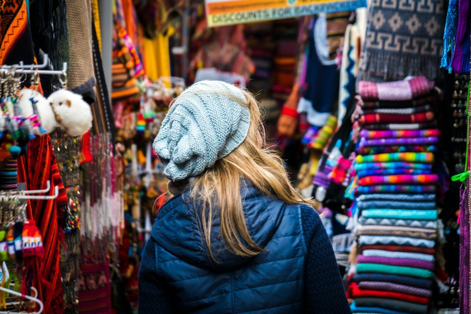 A woman with a knit cap explores a colorful outdoor market full of textiles and crafts.