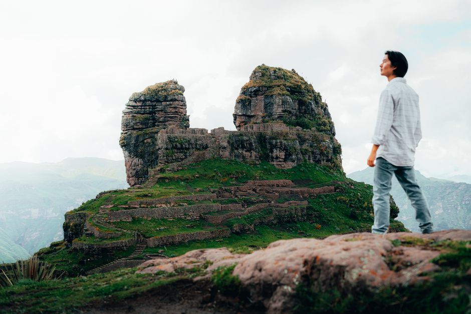 A man stands gazing at the ancient rock formation of Waqrapukara in Cusco, Peru, surrounded by lush mountains.