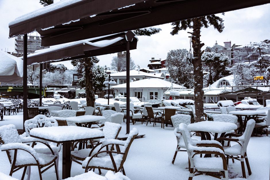 A winter scene of an outdoor restaurant covered in snow, with empty chairs and tables.